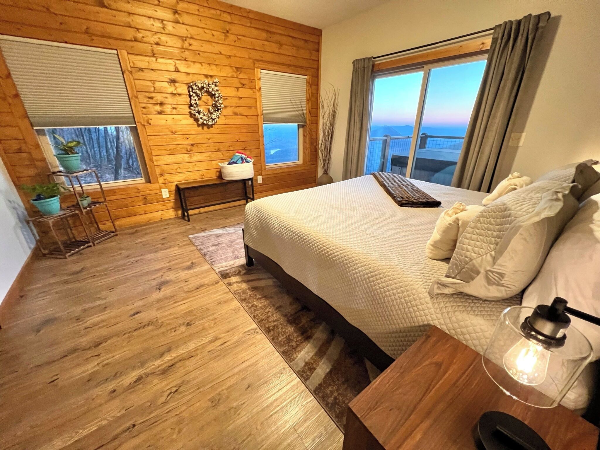 A bedroom with a wooden floor, a large bed with pillows and a blanket, a bedside table with a lamp, and a window with a view.