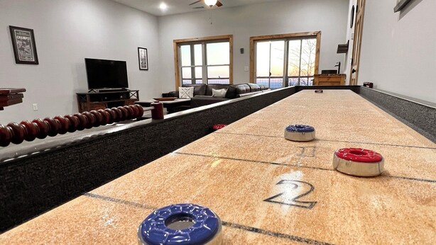 Et shuffleboard-bord med nummererede felter, et fjernsyn og en loftventilator i et rum med store vinduer.