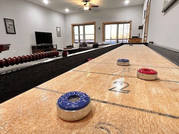 A shuffleboard table with numbered spots, a television, and a ceiling fan in a room with large windows.