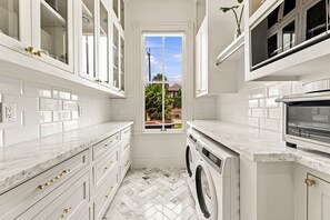 A compact kitchen with white cabinetry, a built-in washing machine, and a window providing a view of the outdoors. - Historic Galveston Cottage | Walk to the Beach (Galveston)