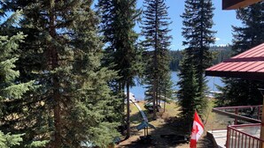 Una terraza de madera con una bandera canadiense, con vista a un lago rodeado de árboles y una cabaña al fondo.