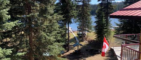 A wooden deck with a Canadian flag, overlooking a lake with trees and a cabin in the background.