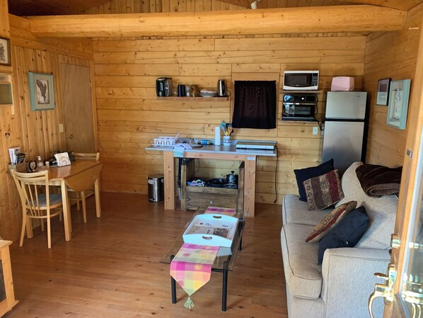 A cozy cabin interior with a dining table, a sofa, a small kitchen area with a microwave and toaster, and a trash bin.