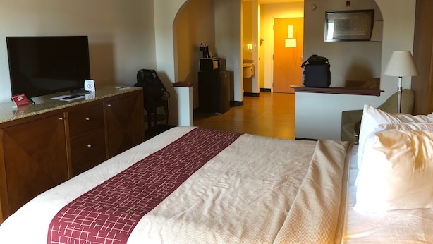 Quarto de hotel com cama, escrivaninha, televisão e banheiro visível por uma arquibancada.