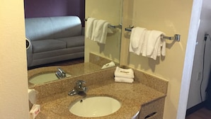 Casa de banho de hotel com lavatório, espelho, porta-toalhas e parte visível de uma cama.