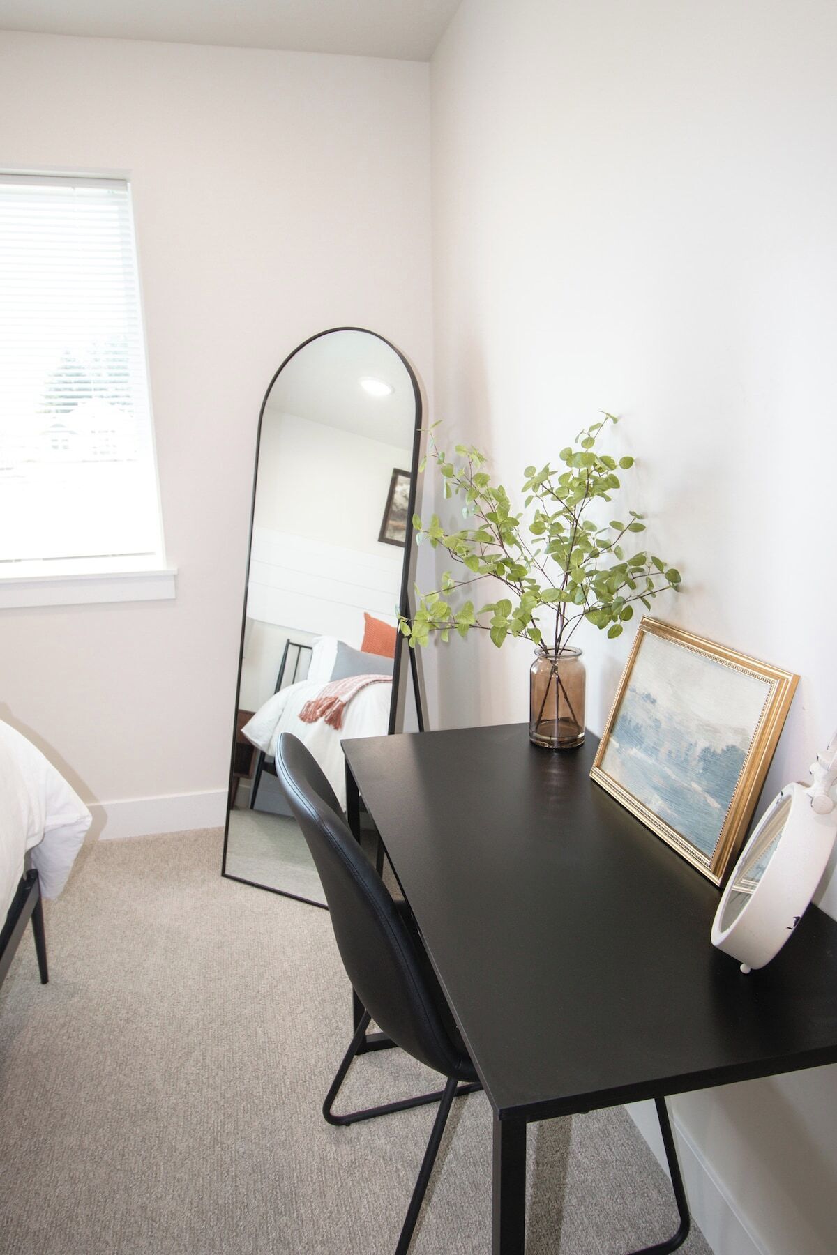 Una habitación con una cama, una mesa con un florero y un marco de fotos, un espejo y una ventana con persianas.