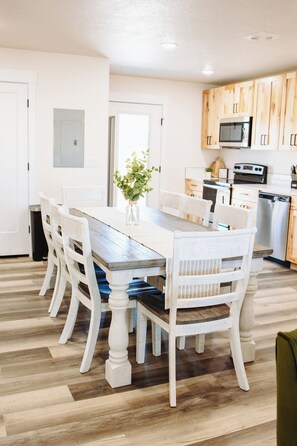 A dining area with a wooden table and chairs, a kitchen in the background, and a white door.