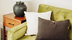 Un canapé vert avec deux coussins, une petite table d’appoint en bois avec une lampe, et un tableau encadré accroché au mur.