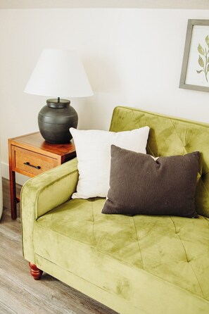 Un canapé vert avec deux coussins, une petite table d’appoint en bois avec une lampe, et un tableau encadré accroché au mur.