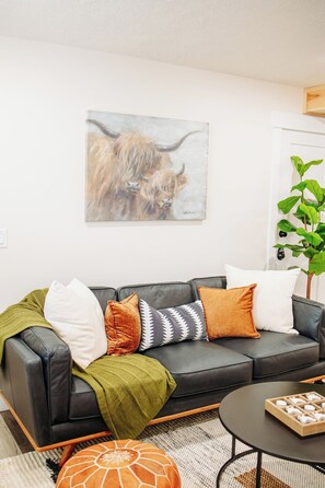 Ein Wohnzimmer mit einem schwarzen Ledersofa, mehreren Kissen, einer grünen Tagesdecke, einem runden Couchtisch mit einer Holzschale und einem Gemälde eines Jaks an der Wand.