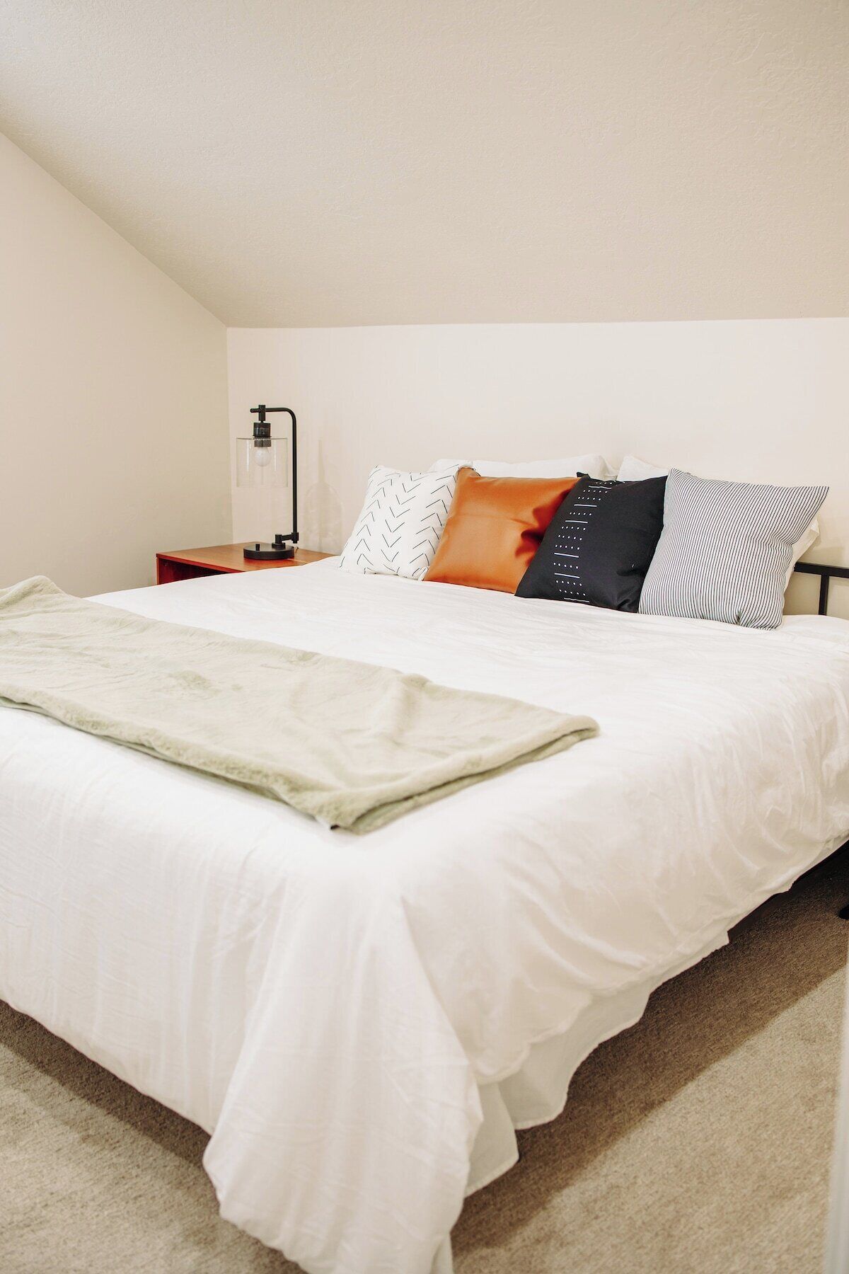 A neatly made bed with white bedding and a throw blanket, a bedside table with a lamp, and a wall-mounted shelf.