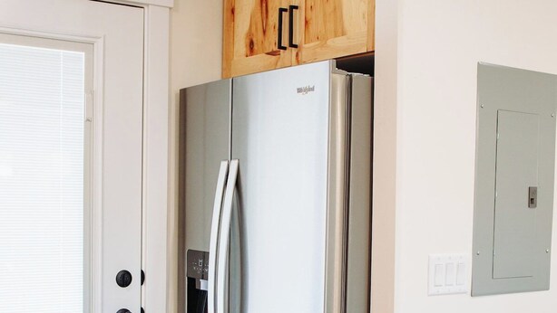 Una cocina con un refrigerador de acero inoxidable, un armario de madera encima y un bote de basura a la derecha.