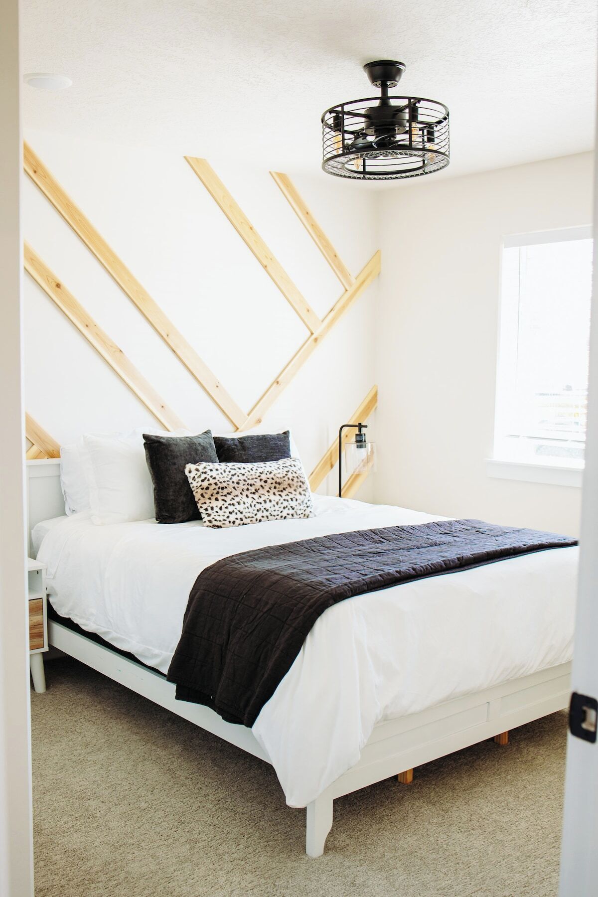 A bedroom with a bed, pillows, a bedside table, a nightstand, a wall-mounted lamp, and a window.