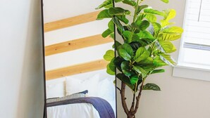 Ein Bett mit weißblauer Decke, ein Spiegel mit Holzrahmen und ein Blumentopf mit grünen Blättern.