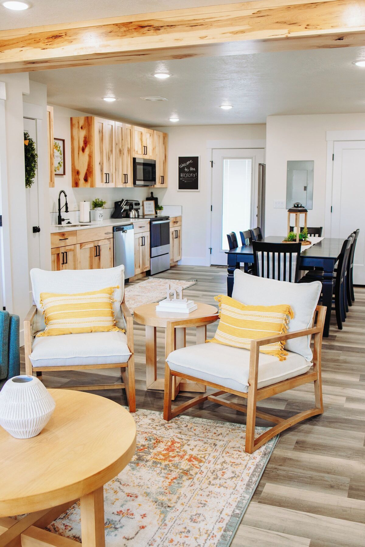 A modern kitchen with wooden cabinets, a dining area with chairs and a table, and a living space with sofas and a rug.