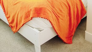 Ein Einzelbett mit weißem Gestell, einer weißen Matratze und einer Decke, die von Orange zu Gelb übergeht. Zwei Kissen, eines blau und eines gemustert.