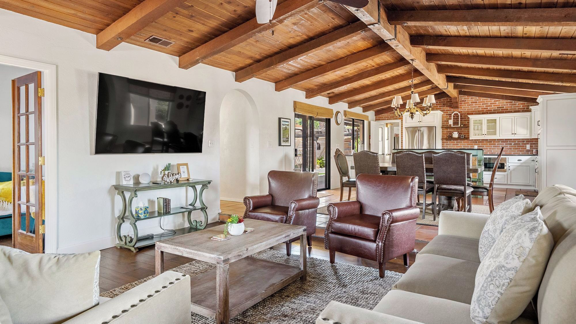 A spacious living room with a wooden ceiling, a television mounted on the wall, leather armchairs, a sofa, and a dining area with a brick wall and white cabinets.