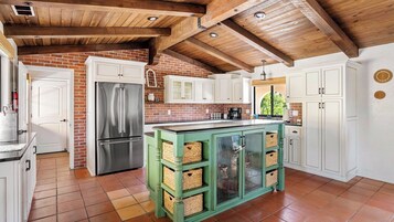 Una cocina con pared de ladrillo, vigas de madera en el techo, armarios blancos, un refrigerador de acero inoxidable e isla verde con cestas.