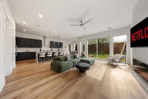 A modern living space with a kitchen, dining area, and a large TV screen. - Modern Trinity Groves Home w/ Rooftop + Garage (Dallas)