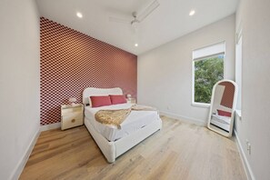 A bedroom with a checkered wall, a bed, a nightstand, a bedside lamp, and a window with a view of trees. - Modern Trinity Groves Home w/ Rooftop + Garage (Dallas)