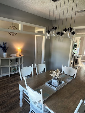 Un espace repas comprenant une longue table en bois, des chaises blanches, un luminaire suspendu et une petite table d’appoint avec une lampe et des objets décoratifs.