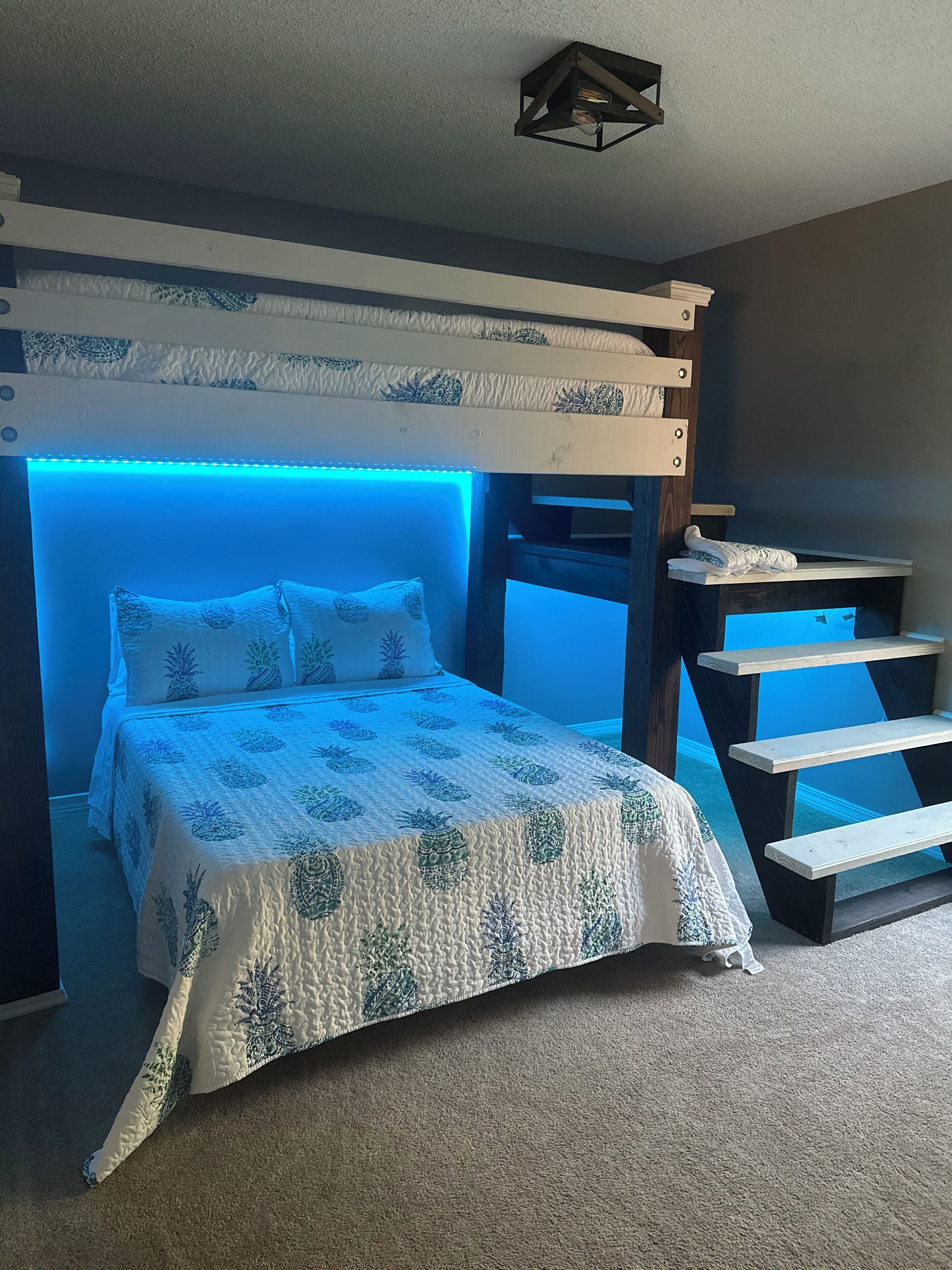 A bunk bed with a headboard illuminated by blue LED lights, a wooden bench, and a patterned bedspread.