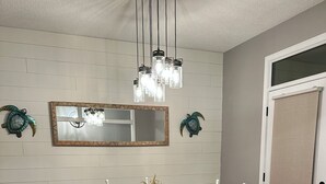 A dining area with a wooden table and white chairs, a hanging light fixture, and a mirror on the wall. - Coastal  Serenity 3-bedroom house with heated pool and hot tub (Gulfport)