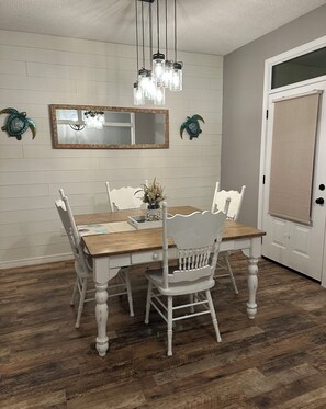 A dining area with a wooden table and white chairs, a hanging light fixture, and a mirror on the wall. - Coastal  Serenity 3-bedroom house with heated pool and hot tub (Gulfport)