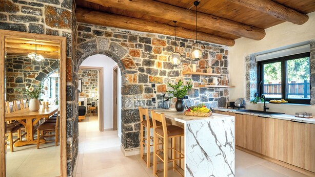 A stone-walled kitchen with a marble island, wooden chairs, and a view of the outdoors.