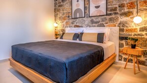 A bedroom with a stone wall, a bed with a dark blue bedspread, two framed artworks, and a small wooden side table with a plant.