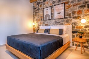 A bedroom with a stone wall, a bed with a dark blue bedspread, two framed artworks, and a small wooden side table with a plant. - Stone horizon villa (Myrthianos Plakias)