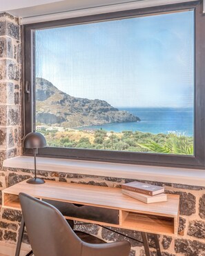 A room with a large window overlooking a coastal landscape, a desk with a chair, and a book titled 'Curated Corners'. - Stone horizon villa (Myrthianos Plakias)