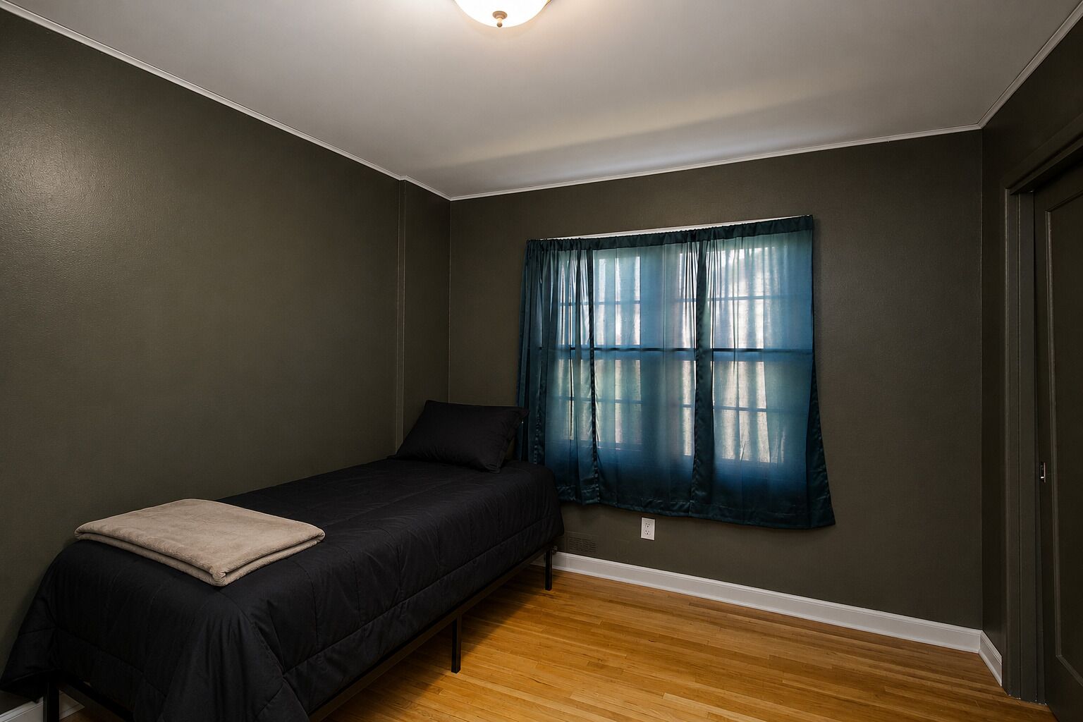 Um quarto pequeno com uma cama de solteiro, piso de madeira, uma janela com cortinas azuis e um lustre no teto.