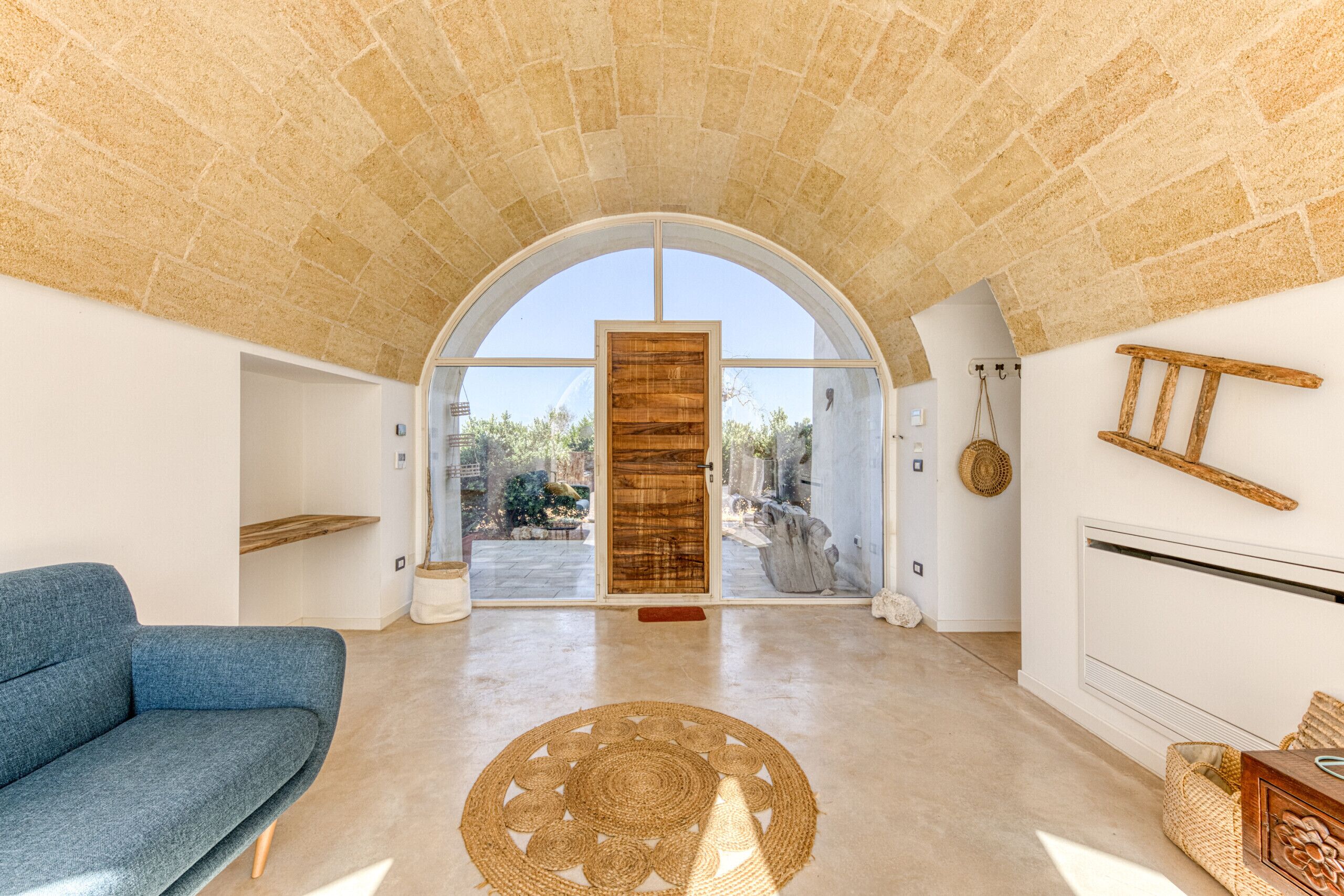 Un'ampia zona giorno con un divano blu, un tappeto tessuto e un arco in pietra che conduce a una porta in vetro.