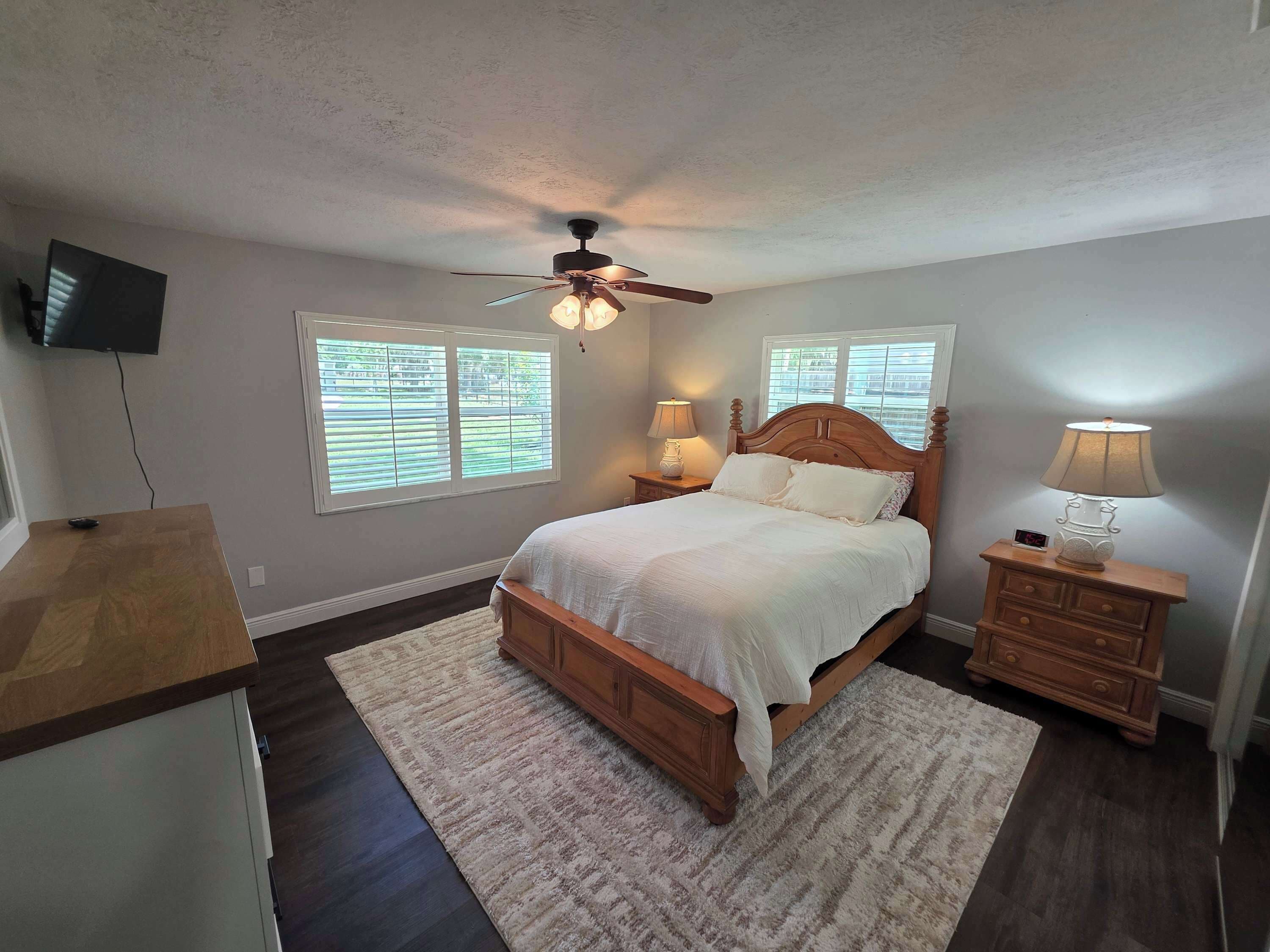 A bedroom with a bed, two bedside tables with lamps, a ceiling fan, and a TV mounted on the wall.