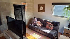 A living room with a sofa, a flat-screen TV, a brick wall, a window with a plant, and a decorative wooden piece on the wall. - Beachwood Villa - Modern Townhouse Walk to Beach (Crescent Head)