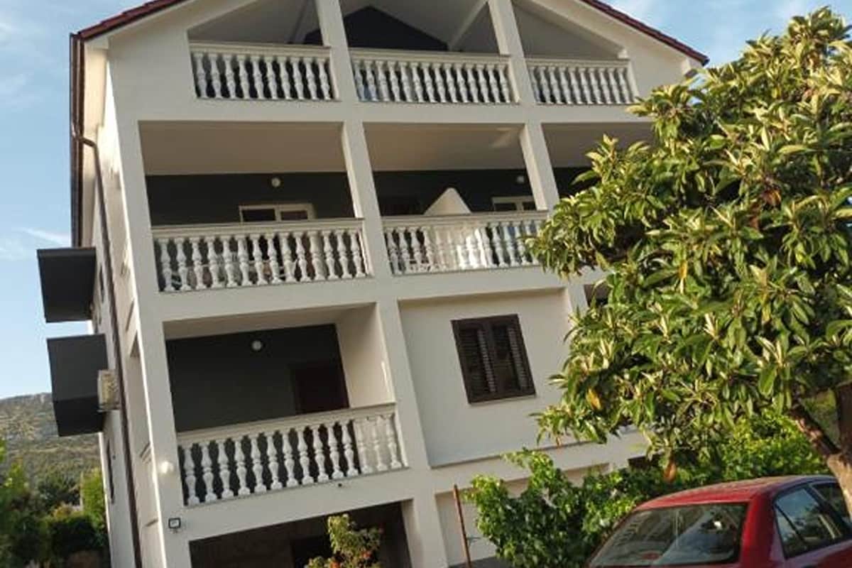 A multi-story building with balconies, a red car parked in front, and a tree to the right.