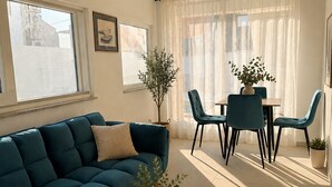 A living room with a blue sofa, a coffee table with a vase and candle, a dining table with chairs, and a large window with sheer curtains. - Apartment Karma (Okrug)