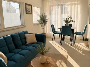 A living room with a blue sofa, a coffee table with a vase and candle, a dining table with chairs, and a large window with sheer curtains. - Apartment Karma (Okrug)