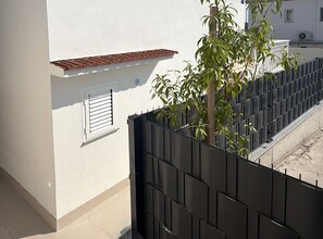 A white building with a black metal fence, a small window with shutters, and a tree in front of the fence. - Apartment Karma (Okrug)