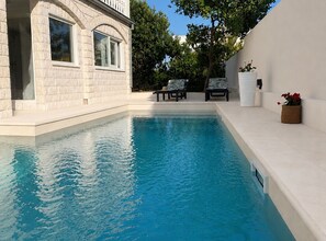 A swimming pool with a clear blue water surface, surrounded by a white building with large windows and a balcony. There are two lounge chairs and a potted plant on the poolside. - Apartment Karma (Okrug)