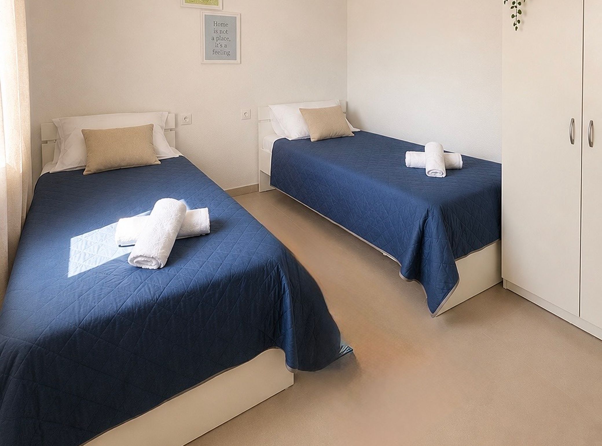 A hotel room with two single beds, each with a blue bedspread and white pillows, and a pair of rolled towels on each bed.
