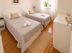 A hotel room with two beds, a small bedside table with a lamp, a wooden floor, and a window with curtains. - Apartment Karma (Okrug)