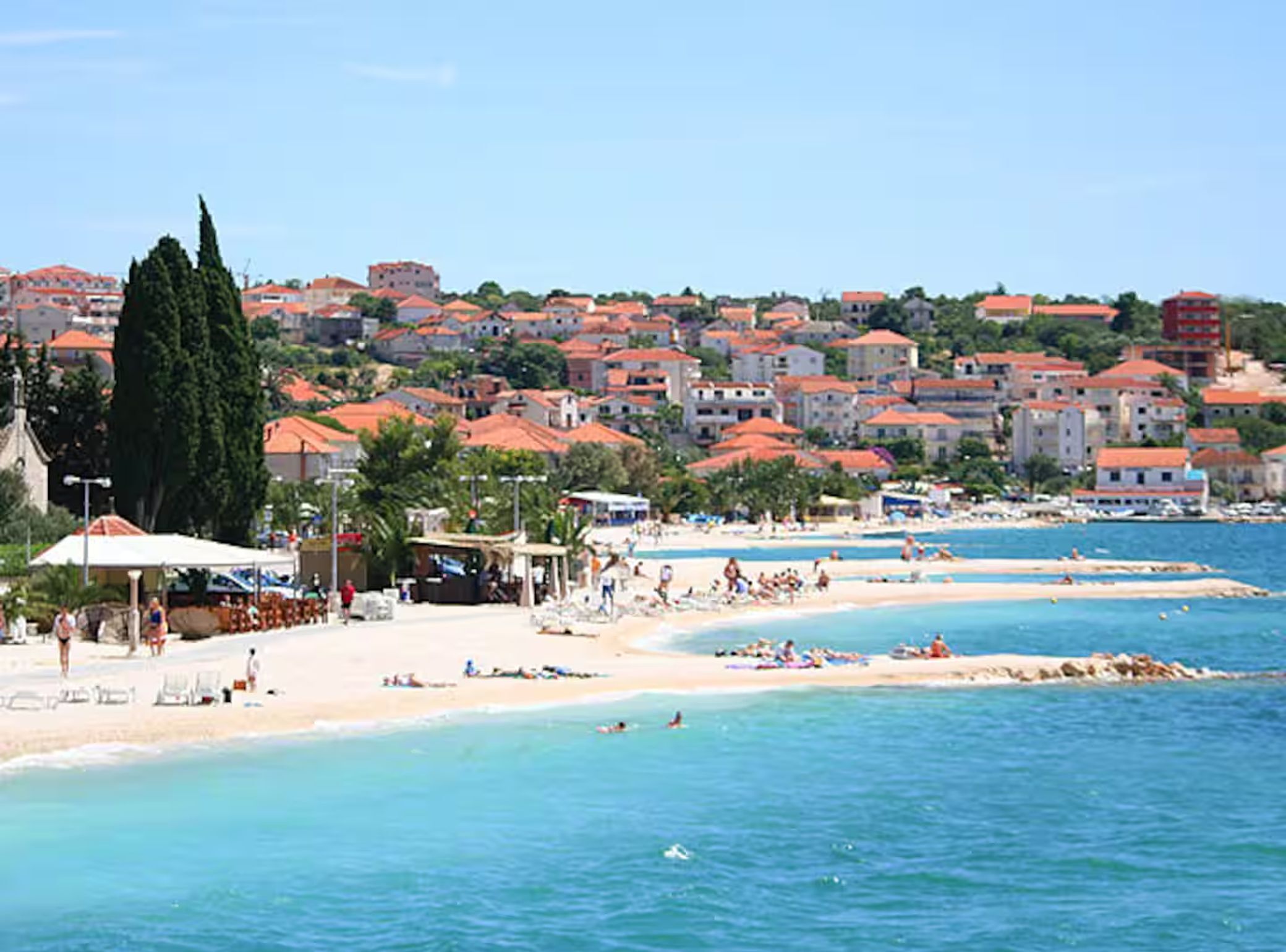 A coastal town with a beach, clear blue waters, and buildings with red roofs.