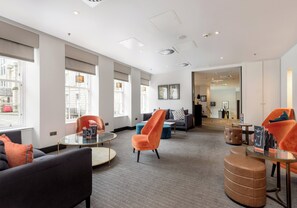 Eine moderne Hotelhalle mit einem runden Glastisch, orangefarbenen und schwarzen Stühlen und Blick auf die Straße durch große Fenster.