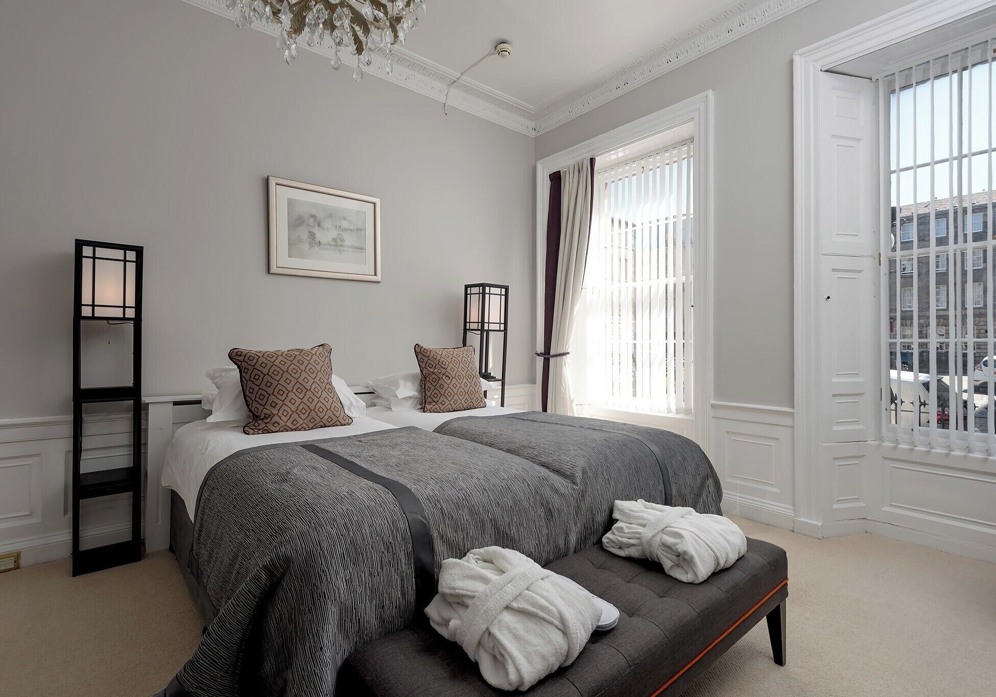 A bedroom with a large bed, two bedside tables, a chandelier, a window with curtains, and a view of the outside.