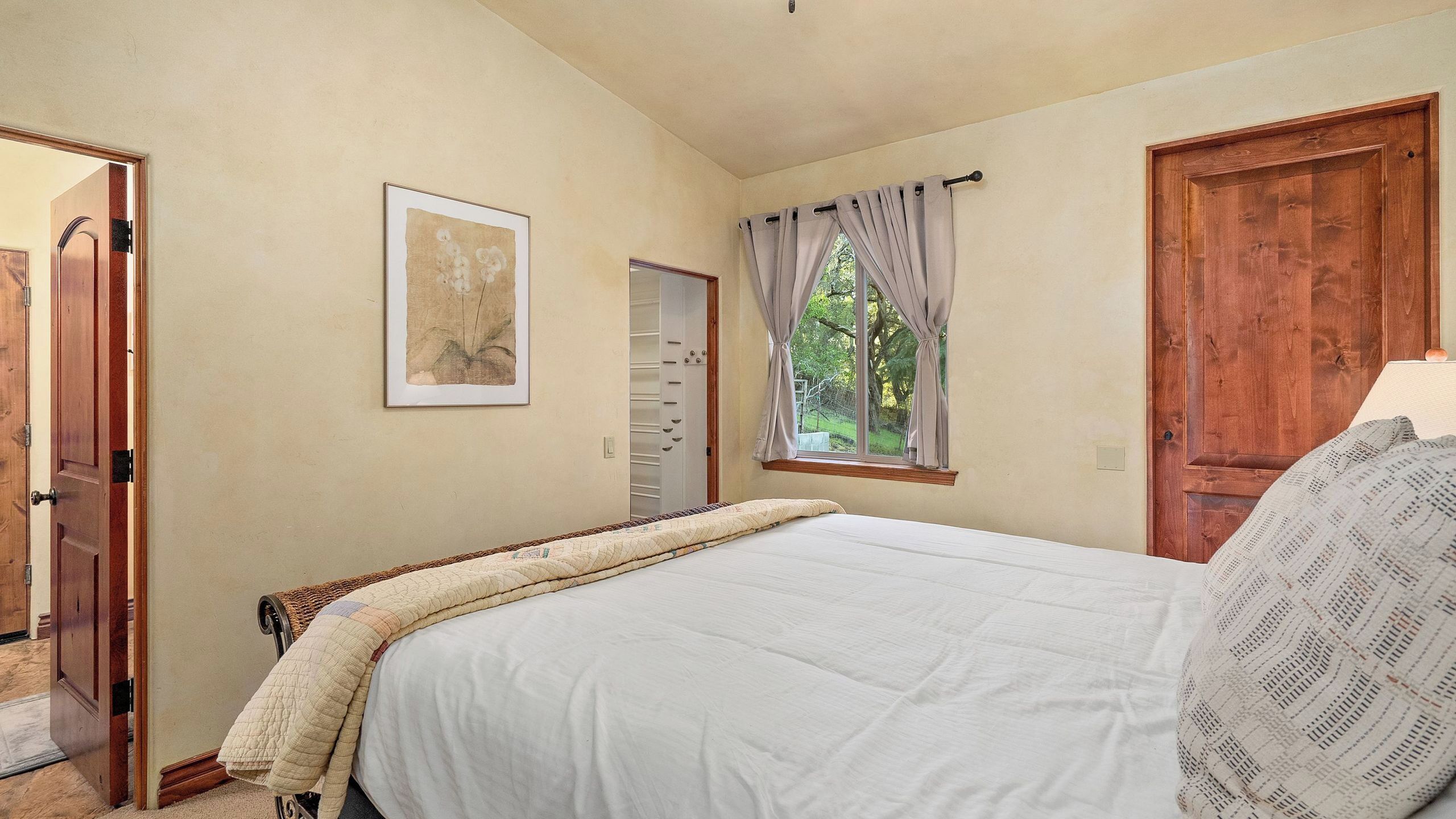 Un dormitorio con cama, ventana con cortinas, puerta y un cuadro en la pared.