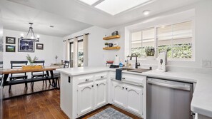 A modern kitchen with white cabinetry, a built-in dishwasher, and a window with a view of greenery. - Whispering Wine by Avantstay Country Acreage + Fire Pit + Deck Central Coast Wine Country (Paso Robles)