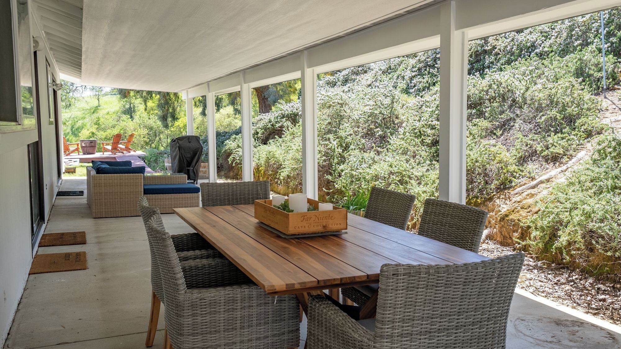Una veranda esterna con tavolo in legno e sedie in vimini, circondata da ampie finestre con vista su un rigoglioso giardino.