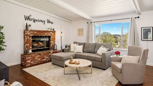 A living room with a fireplace, a grey sofa, a round coffee table, and a view of the outdoors. - Whispering Wine by Avantstay Country Acreage + Fire Pit + Deck Central Coast Wine Country (Paso Robles)